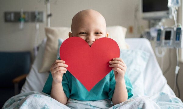 Bald Caucasion Child with red paper heart in hospital bed. Concept of health, hope, recovery for Valentine's Day or childhood cancer with copy space.