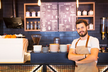 The barista, wearing a white t-shirt and beige apron, has his arms crossed and is confident. Various coffee cups and a coffee maker are visible on the counter.