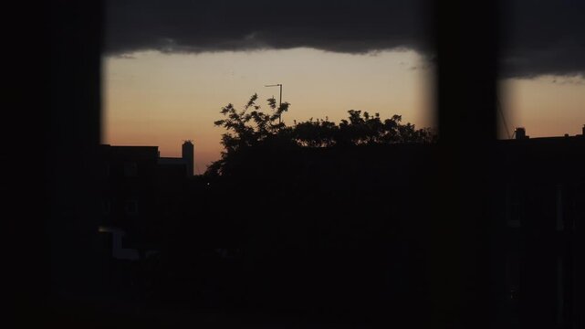 Footage captured from indoors through a window, showing a deep red sunset sky above urban surroundings in London, England. The scene features city silhouettes, warm evening colors, and atmospheric lig