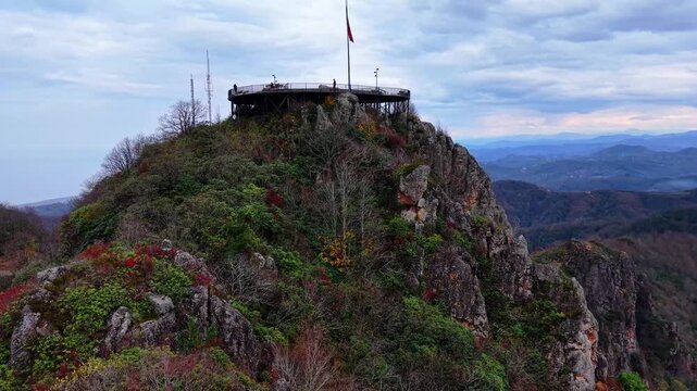 A cinematic drone shot flying between sharp rocky peaks toward a scenic mountain observation terrace. Colorful autumn trees in vibrant red, orange, and yellow tones cover the landscape, creating a bre
