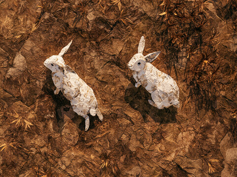 Two rabbits made of paper sit on brown leaves during a sunny afternoon in a forest setting