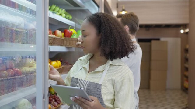 Grocery store worker and woman choosing fresh fruit in produce aisle with tablet and apron near refrigerated display for healthy food selection and shopping search optimization