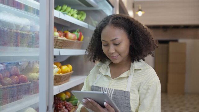 Young woman shopping grocery store aisle, selecting fresh fruit with tablet, female worker grocery store choosing fruit cold display, holding tablet and checking shelf stock, grocery store worker