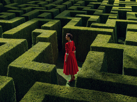 Woman in red dress stands in large green maze during bright daylight exploring pathways and choices ahead in the garden