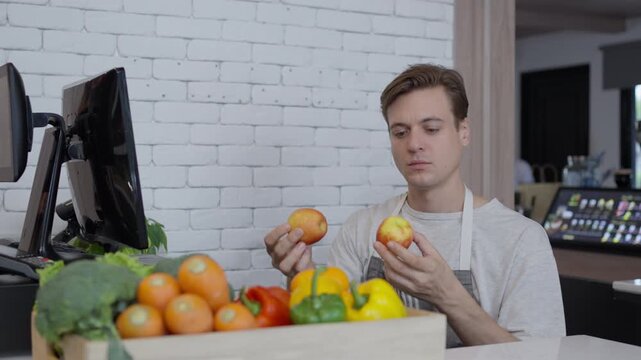 Young man comparing fresh apple and fruit produce at grocery store market checkout, sorting vegetables at indoor shopping counter with apron and counter display