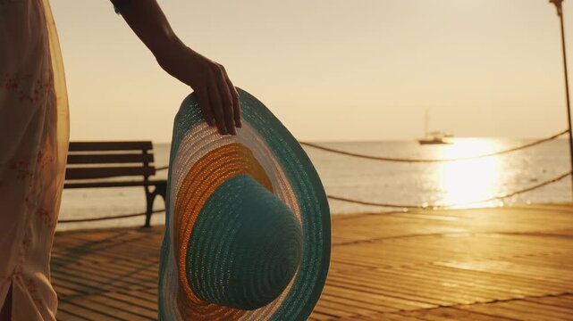 Woman walking on wooden pier with straw hat in hand, scenic sea view at golden hour, peaceful summer evening.
