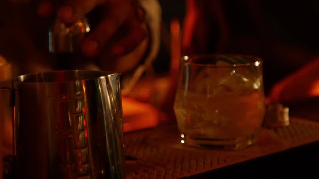 Bartender pouring liquor from jigger into cocktail shaker tin at bar counter with warm amber nightlife lighting