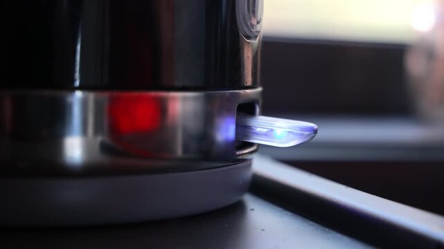 Finger Turning On Electric Kettle Switch In Kitchen, close up home video frame with red indicator light and soft window glow