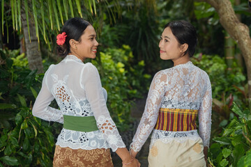 Obraz premium Two Balinese women in white lace kebaya holding hands and smiling at each other in tropical garden