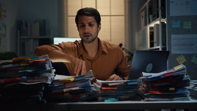 Overworked young Latin businessman working late on document in large stack of paperwork files at office night. Tax audit deadline, accounting stress, corporate burnout, urgent business concept.