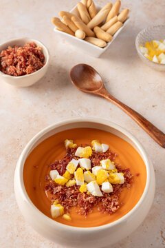 Vertical view of beige bowl with traditional Andalusian salmorejo topped with boiled egg and serrano ham, with ingredient bowls, breadsticks and wooden spoon on light stone background with copy space