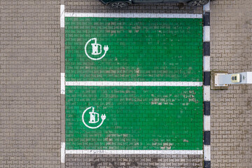Obraz premium Car charging parking lot. Top-down aerial view of two green eco-friendly electric vehicle charging parking spaces with white painted battery plug symbols on interlocking concrete pavers. Electric cars
