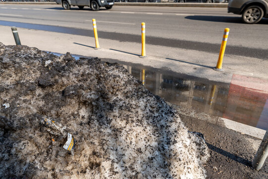 A large melting pile of dirty snow mixed with mud and street debris on the side of a city asphalt road with yellow traffic delineator posts in the background. Dirt snow in the city.