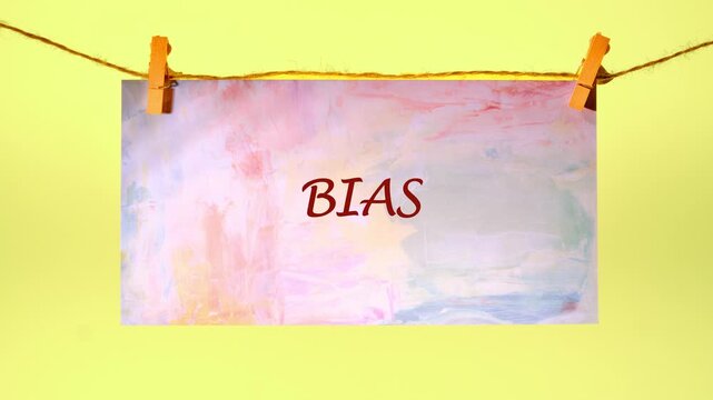 A hand writes the word BIAS with a marker on a pastel watercolor paper. The finished sign hangs on a clothesline with wooden clothespins against a vibrant yellow wall