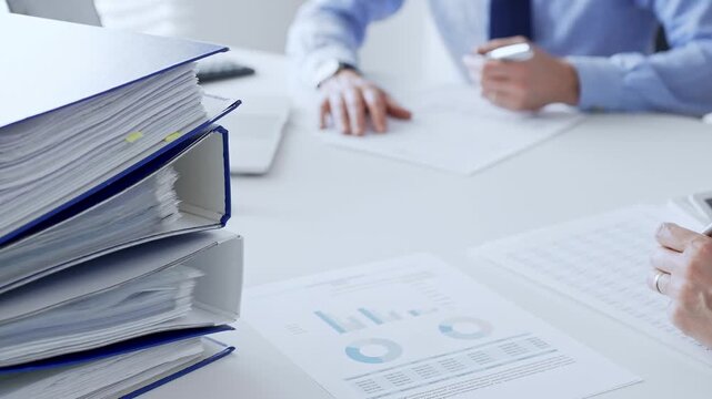 Office workers or auditors are analyzing financial reports and checking documents while sitting at white table with a stack of blue folders in the foreground. Audit and taxes theme in business