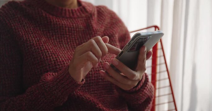 Hand. Asian woman using smartphone on chair by window, scrolling screen with finger in bright home interior, mobile technology and digital lifestyle concept