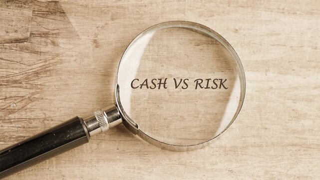 Magnifying glass slides across a light wooden table surface, bringing the etched words Cash Versus Risk into clear focus for financial insights