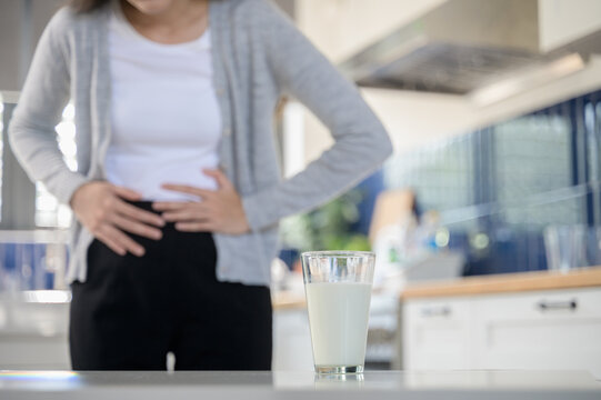 Allergy dairy food lactose intolerance allergen health care illness concept. Close up a glass of milk with blurred Asian woman holding stomach belly feel pain digestive unhappy after drink a cow milk.