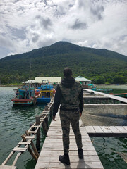 Fototapeta premium Man in camouflage standing on wooden pier overlooking water and fishing boats