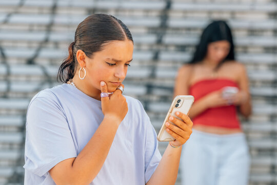 young woman looks at her phone with a thoughtful expression. A second woman stands behind her. Both hold phones outdoors near bleachers