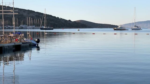 A calm morning at a Bitez marina in Bodrum. A wooden pier with empty restaurant tables stands over glass-like water reflecting parked sailboats, yachts, and the serene Turkish coastline.