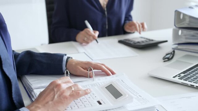 Two professional accountants collaborating in a modern office, calculating taxes and analyzing financial data with calculators, documents, and a laptop. Audit and taxes in business