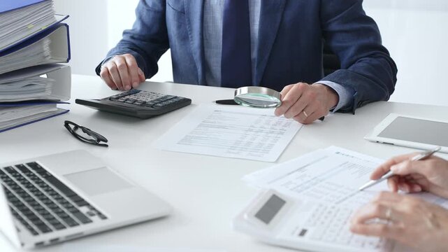 Male financial analysts examining documents, using magnifying glass and calculator while studying corporate data at workplace with a female colleague. Audit and taxes concept