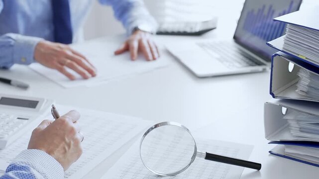 Accountants are analyzing financial reports with magnifying glass and documents, performing accounting and auditing tasks in the office, close up. Audit and taxes in business