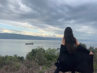 Fototapeta premium Back view of a young woman looking at the sea with a ship from a high mountain viewpoint.