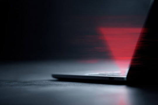 Laptop on office desk showing abstract red warning symbol and glitch effect, cyber attack and data breach concept, dark background, realistic stock photo, no readable text