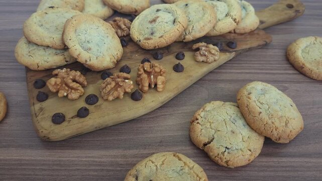 Close-up view of homemade cookies with chocolate chips and walnuts arranged on a rustic wooden board. Freshly baked golden biscuits with a crunchy texture are scattered alongside whole walnuts 