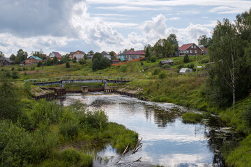 Fototapeta premium Summer rural landscape. View of a dam with a pedestrian bridge over a small river. Village houses in the background. Imeni Babushkina village, Vologda Oblast, Russia. Ledenga River.