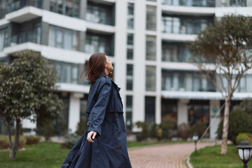 Obraz premium Young woman in a long dark coat enjoys a walk in an urban park with modern buildings in the background during a calm day.