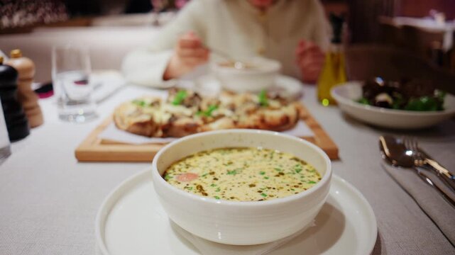 Close up of a bowl of creamy soup served on a restaurant table with pizza, salad, olive oil, and tableware in the background