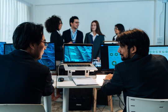 Two professional businessmen discussing digital data on computer screens in a modern office with team in background. Cybersecurity experts and software developers working on corporate tech project
