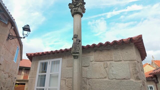 Descending aerial view of a traditional stone cross, known as a cruceiro, revealing the narrow cobblestone street and historic stone buildings of the charming fishing village of Combarro in Galicia