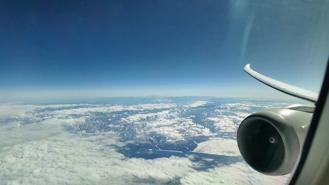 Scenic aerial look at clouds and landscape from a passenger window, featuring the aircraft wing and jet engine on a bright sunny day.