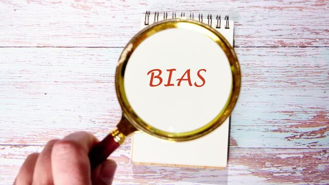 A hand slowly moves a magnifying glass across a white notepad resting on a wooden desk. The glass magnifies barely visible marks into the clear word BIAS