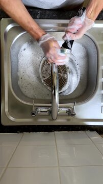 Overhead view of hands cleaning a black skillet pan with a sponge and soapy water in a stainless steel sink