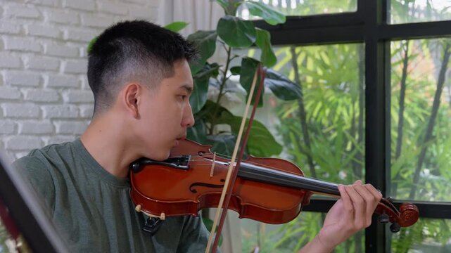 Asian young men practice violin on sofa, their skilled hands guiding the bow across the strings to produce melodious tunes, artistic lifestyle playing violin in living room for practice  and  hobby