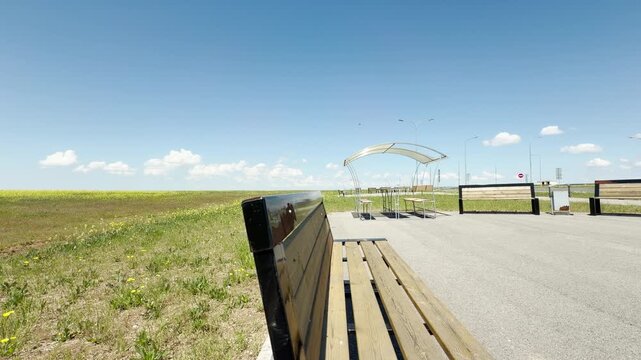 A roadside rest area features wooden benches and a sheltered picnic table next to a grassy field. The vast blue sky and bright sunshine evoke a calm, refreshing, and peaceful travel feeling.