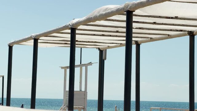 A white fabric sunshade and wooden lifeguard tower sit quietly on a sunny beach. The sparkling turquoise water and light blue sky evoke a peaceful, calm, and deeply relaxing summer mood.