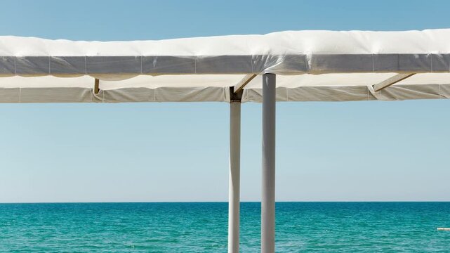 A simple white sun awning frames a stunning view of the vibrant blue sea. The clear sky and sparkling water create a minimalist, deeply peaceful, and refreshing summer mood.