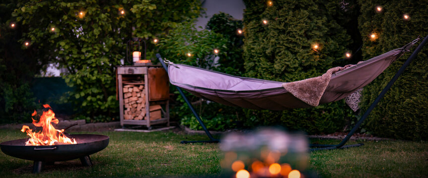Hammock with fairy lights in garden. Cozy evening atmosphere with a campfire in a fire bowl. Background for leisure activities for outdoors.
