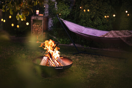 Hammock with fairy lights in garden. Cozy evening atmosphere with a campfire in a fire bowl. Background for leisure activities for outdoors.