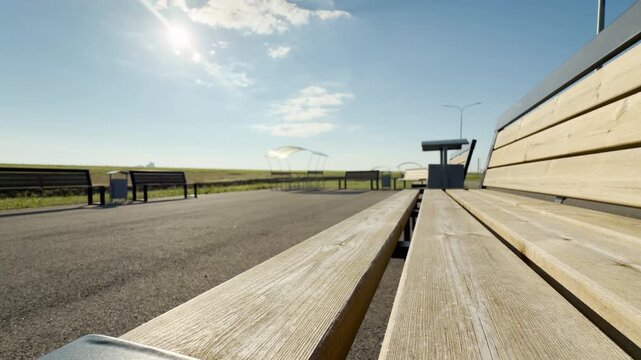 A close-up perspective of wooden benches highlights a peaceful and isolated afternoon at a roadside rest area. The paved path stretches into the distance beneath a warm, bright sky.