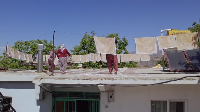 Women dry grape pestil sheets on a rooftop under clear blue sky, showing traditional Turkish fruit leather making, sun-drying, rural harvest work, homemade food preparation and authentic village life.
