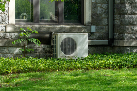 White outdoor air conditioning unit nestled against a vintage stone building with a reflective window, surrounded by lush green grass and vibrant shrubbery on a sunny day.