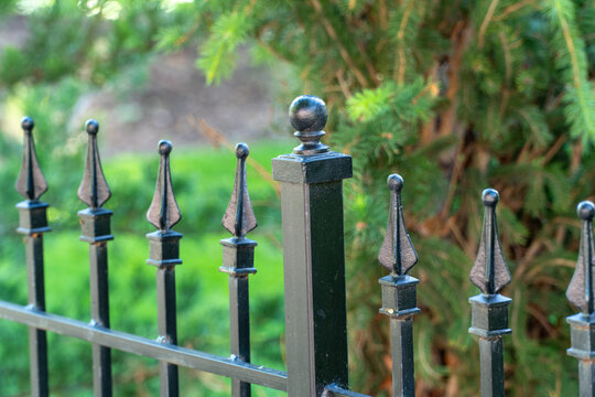 Elegant black wrought iron fence with ornate finials and posts, providing classic security and privacy against a softly blurred vibrant green garden background. Traditional outdoor architecture.