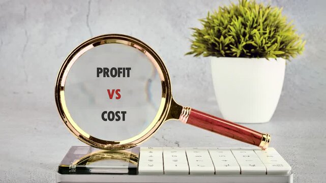 A magnifying glass reveals the words PROFIT VS COST on a white calculator, symbolizing a detailed financial analysis on a gray office desk with a green plant